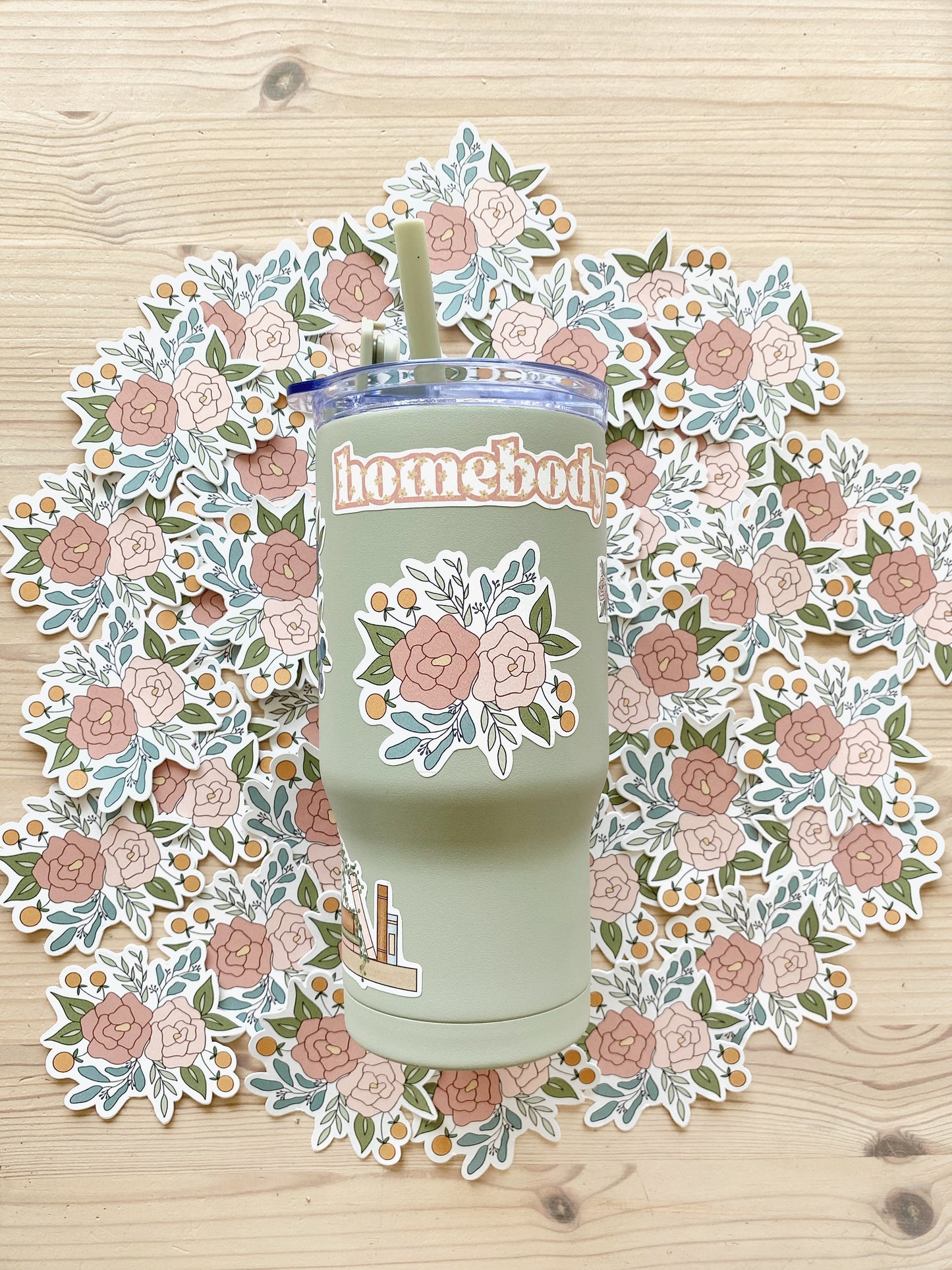 A sage green tumbler featuring a floral arrangement design sits on atop a layer of matching pink peony stickers.