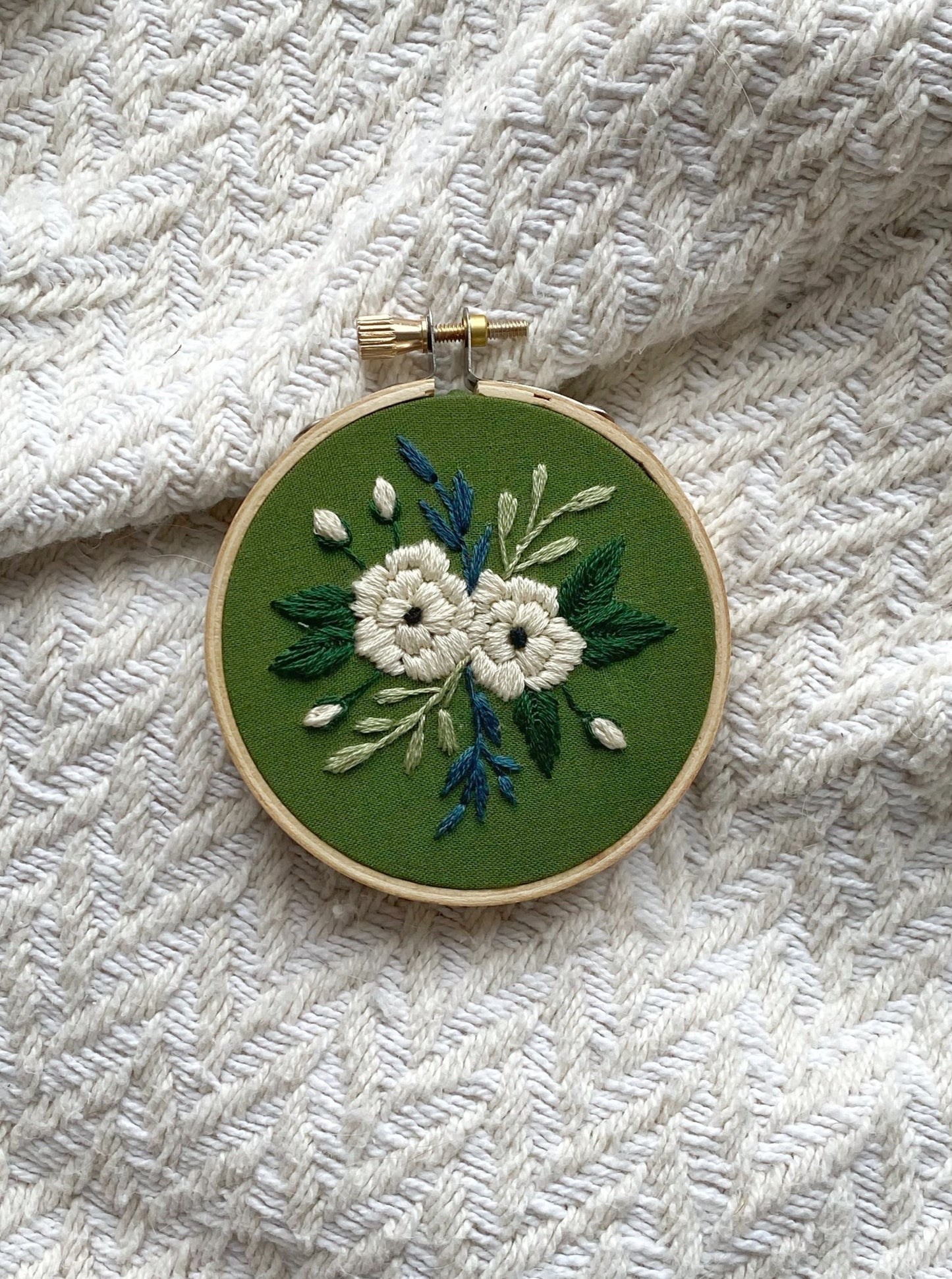 An embroidery hoop art featuring a variety of greenery and small blooms surrounding two large white peonies stitched on deep green fabric and framed in a 3 inch wooden hoop on a textured display background.