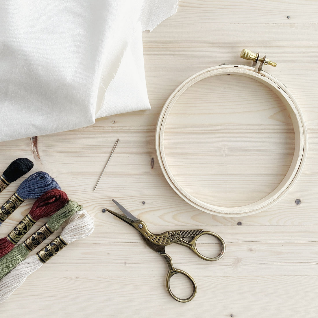Embroidery essentials: a wooden embroidery hoop, an embroidery needle, DMC embroidery floss, quilting cotton fabric, and a pair of stork-shaped embroidery scissors.