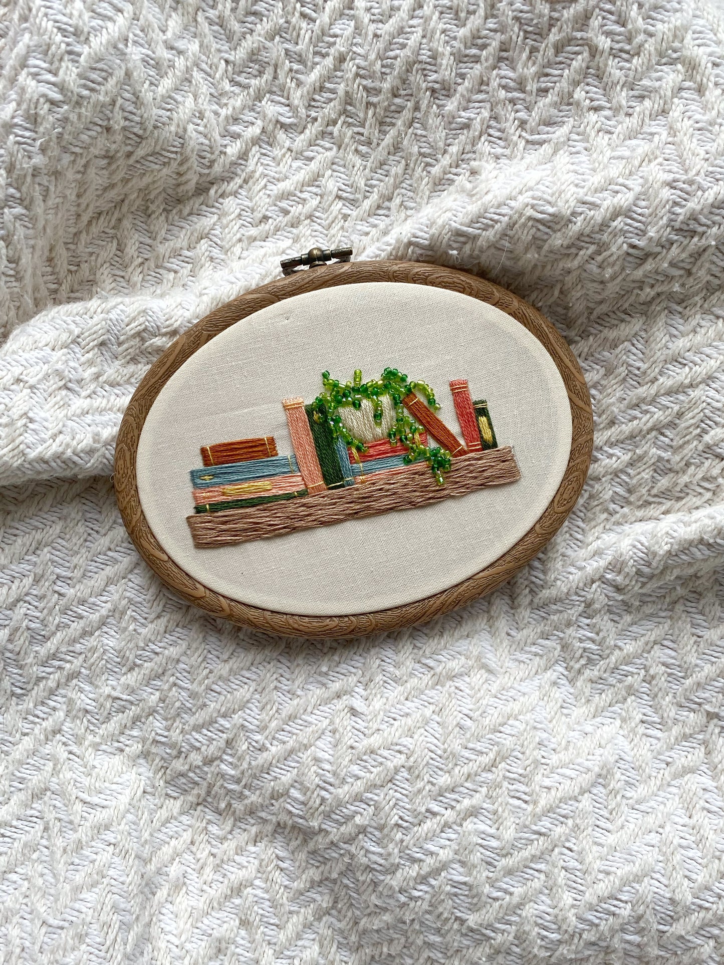 A finished embroidery hoop featuring a shelf with books and a potted plant on a textured surface pattern for beginner embroidery.