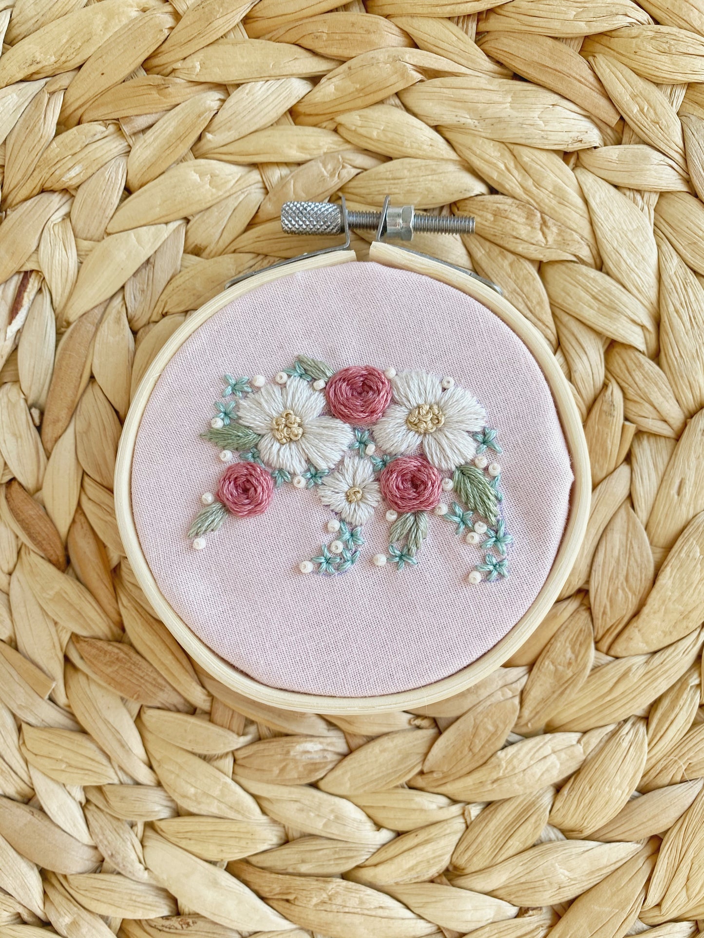 The shape of a bear is embroidered in flowers and framed on pink fabric in an embroidery hoop displayed on a textured background.