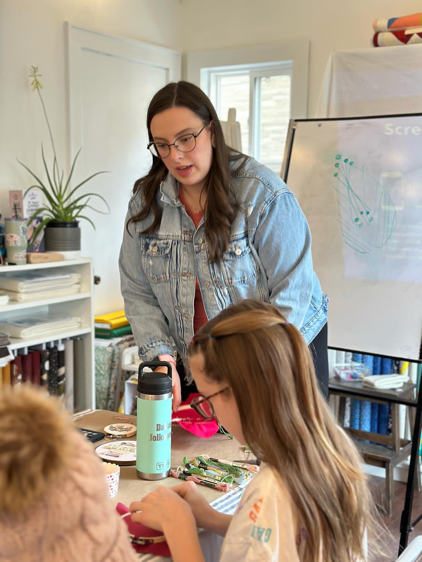 A woman teaching an in person beginner embroidery workshop with a stitch diagram on a whiteboard in the background.