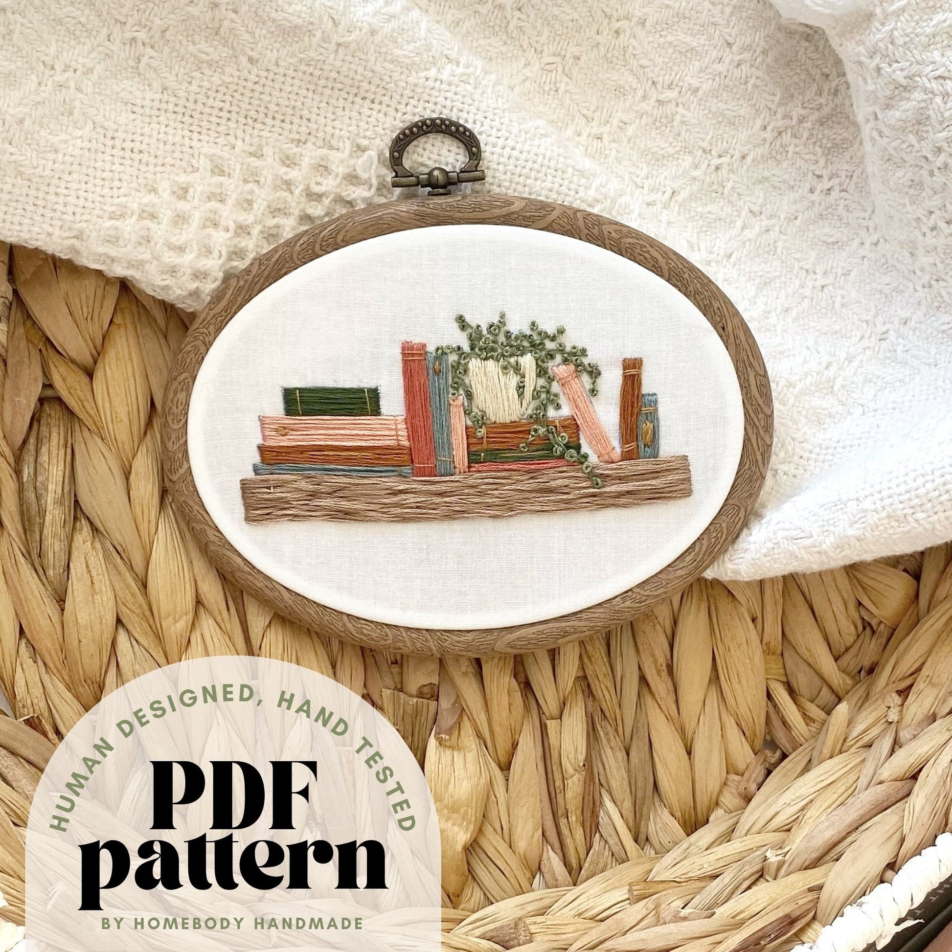 An embroidery hoop featuring a bookshelf design sits on a textured surface. A text overlay in the bottom left corner says "human designed, hand tested PDF pattern by Homebody Handmade."