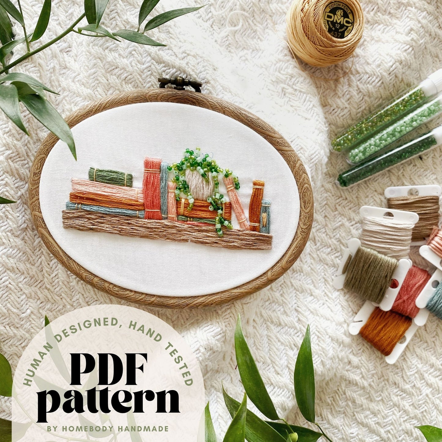 Embroidery hoop with a completed bookshelf design on a textured surface with embroidery supplies around it text overlay indicates hand tested human designed pdf pattern.