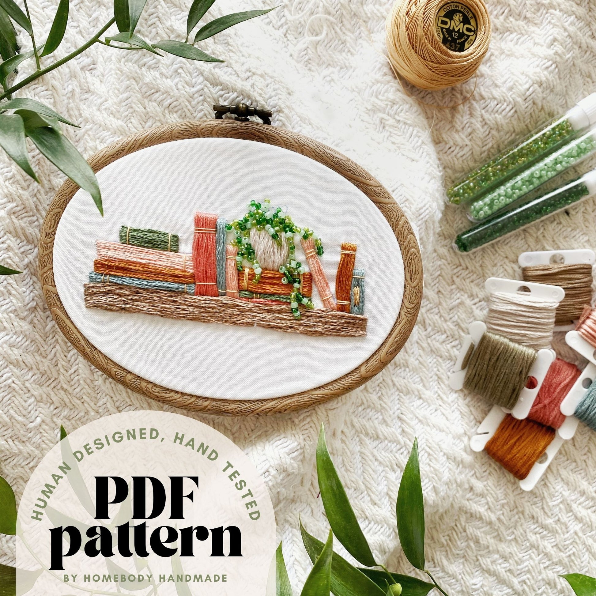Embroidery hoop with a completed bookshelf design on a textured surface with embroidery supplies around it text overlay indicates hand tested human designed pdf pattern.