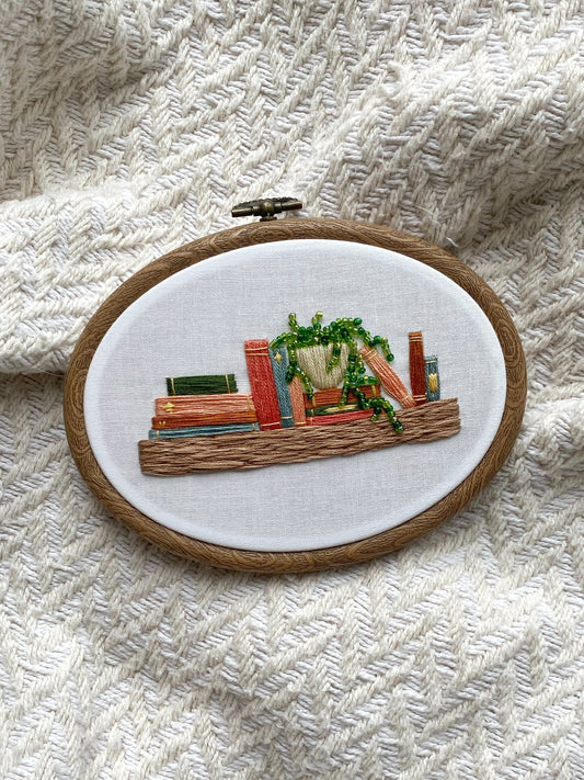 Embroidery hoop artwork depicting a bookshelf with books and a potted plant, stitched in various colors on a textured white background.