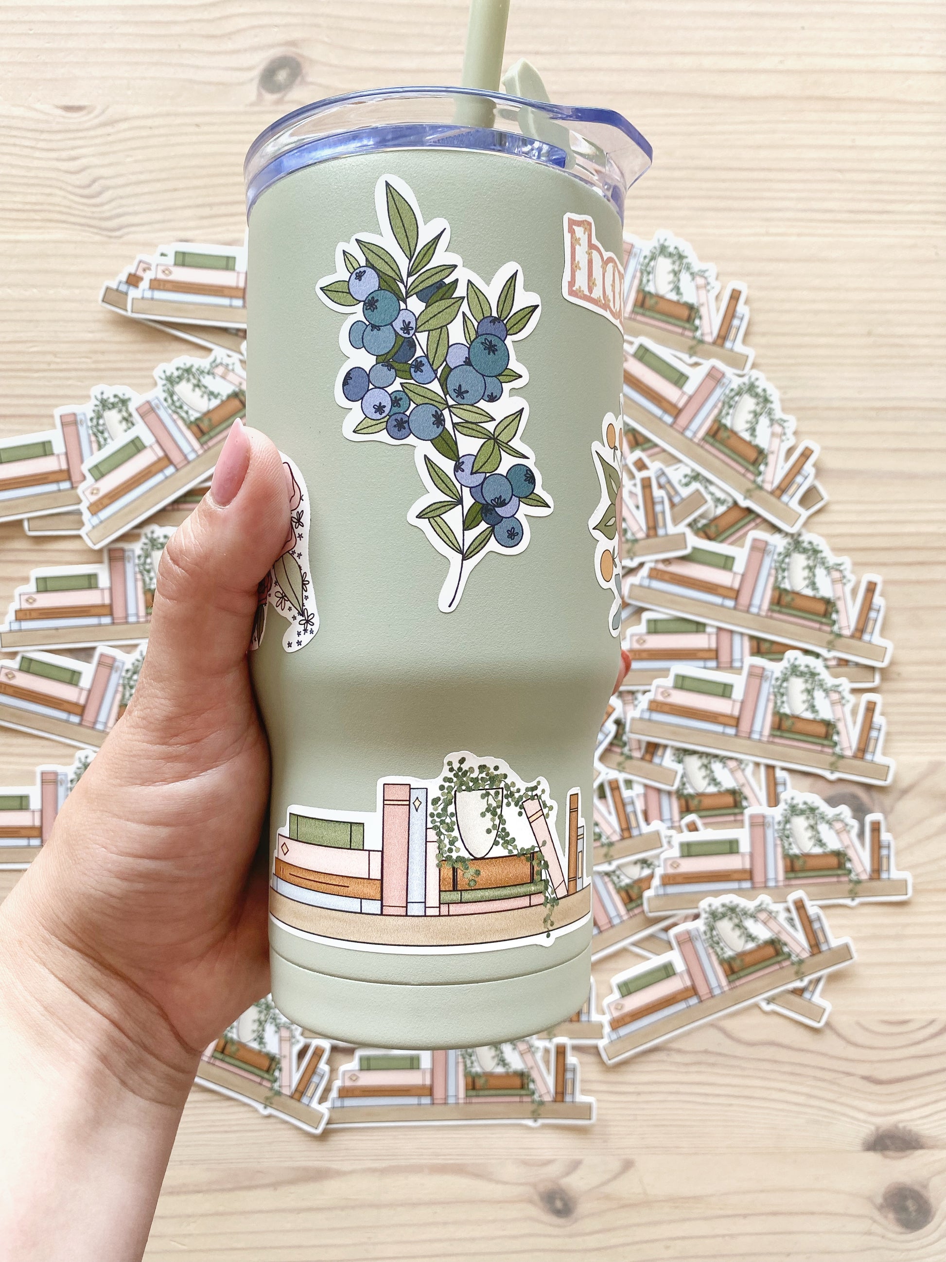 A hand holds a sage green drink tumbler cup with a variety of stickers featured on it including a bookshelf design and wild blueberries the background depicts a variety of matching bookshelf stickers.