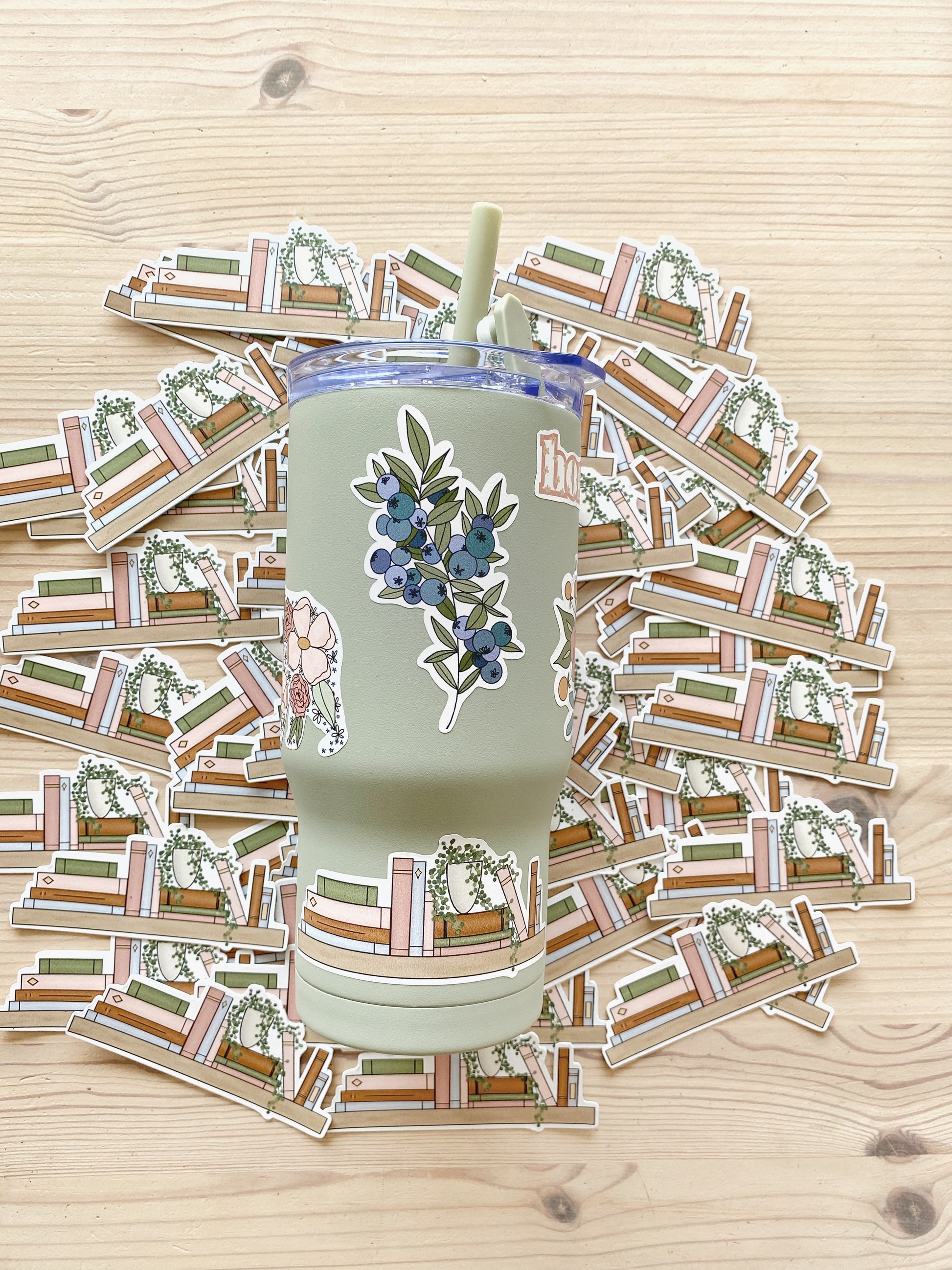 A sage green drink tumbler featuring a variety of illustrated stickers including a sprig of blueberries and a bookshelf design with matching bookshelf stickers in the background.