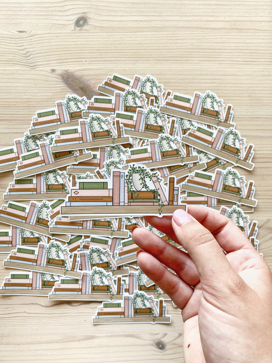 A hand holds a vinyl sticker shaped like a bookshelf featuring a variety of books and a potted plant in front of a background of matching bookshelf stickers.