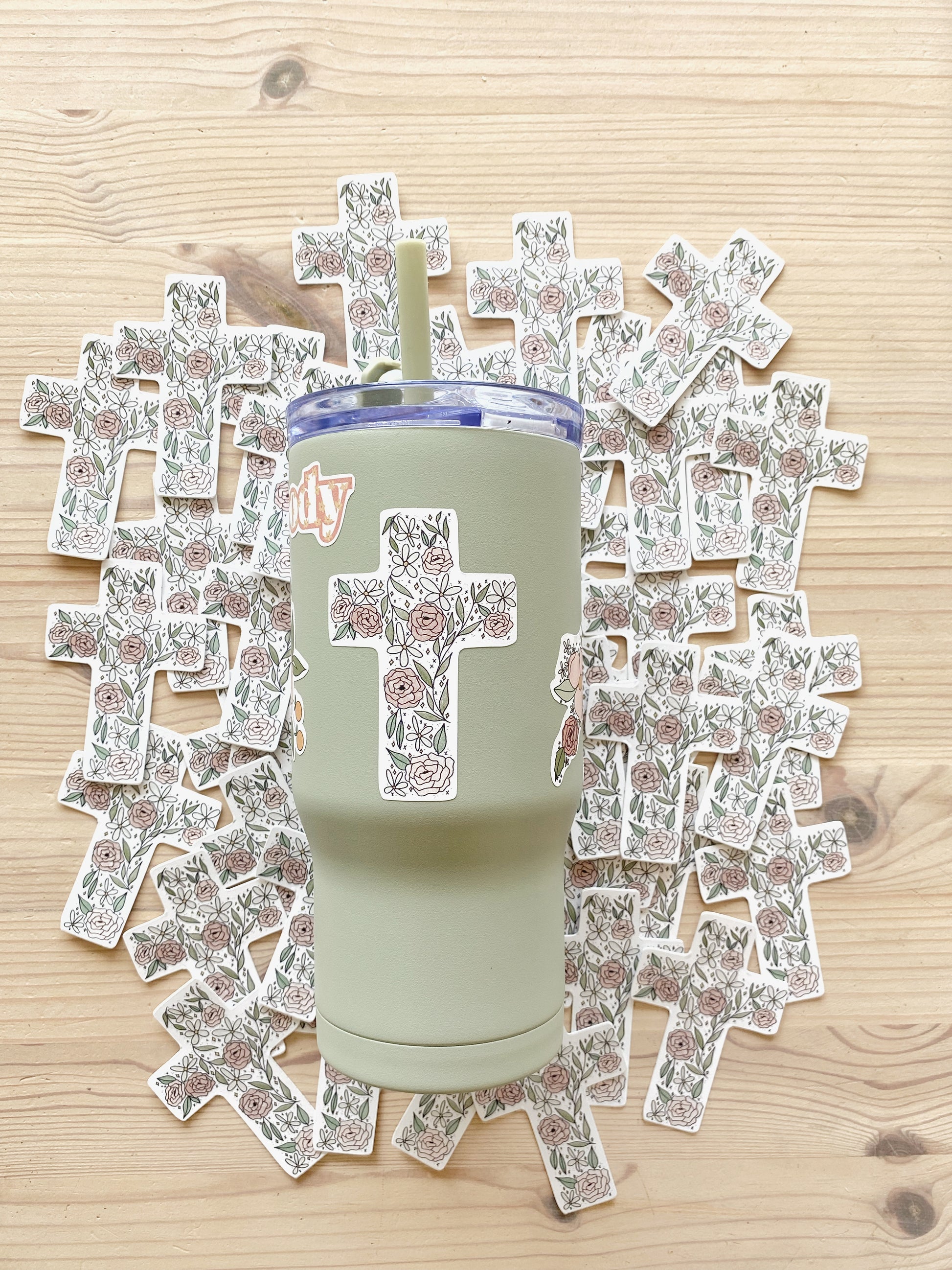 Sage green tumbler cup with floral cross sticker lays atop a background of matching stickers.