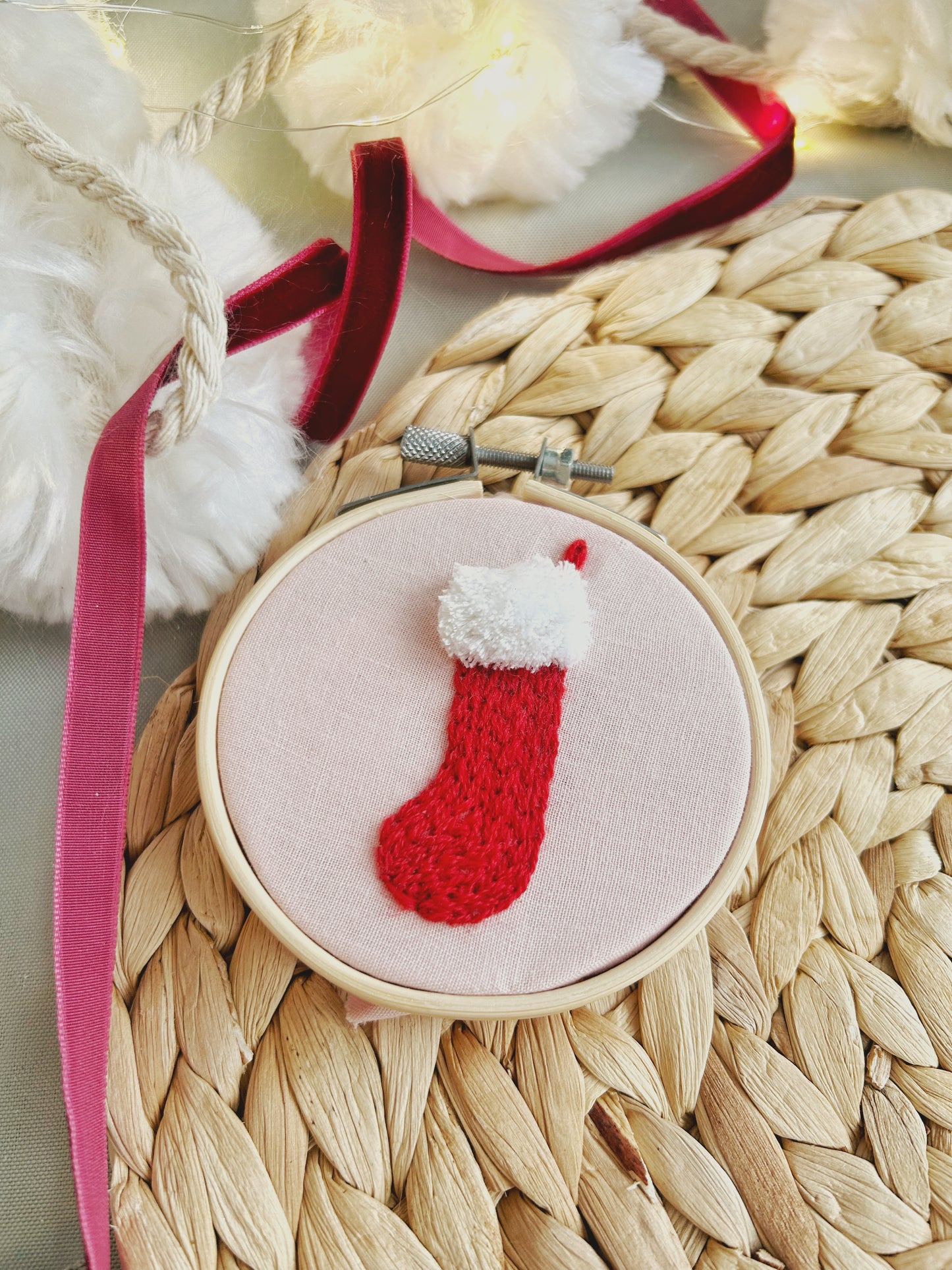 A close up of a Christmas ornament embroidery hoop featuring a knit-look stick and stitch stocking design sits on a textured surface and is surrounded by seasonal decorations.