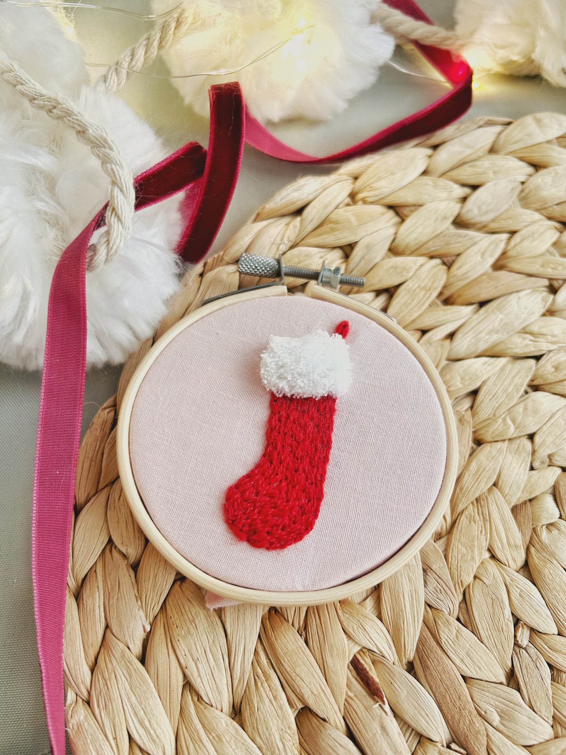 A close up of a Christmas ornament embroidery hoop featuring a knit-look stick and stitch stocking design sits on a textured surface and is surrounded by seasonal decorations.