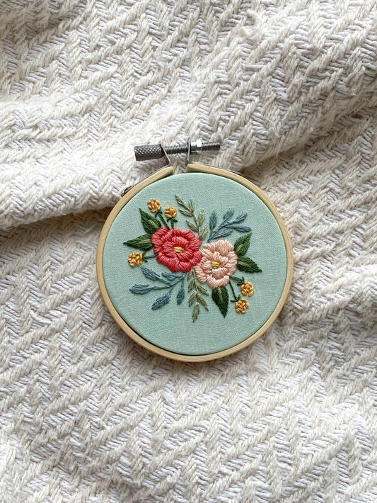 A 3-inch embroidery hoop artwork depicting two peonies and green leaves and foliage on a light blue background, framed in a wooden hoop.