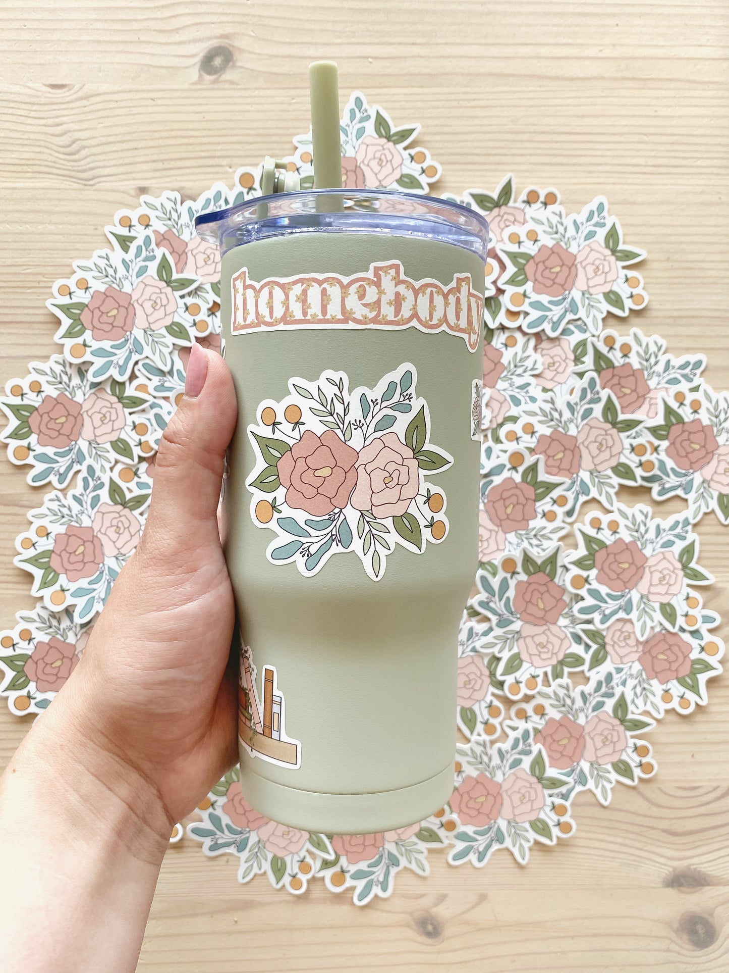 Hand holding a light green tumbler with featuring a floral arrangement sticker with matching floral stickers in the background.