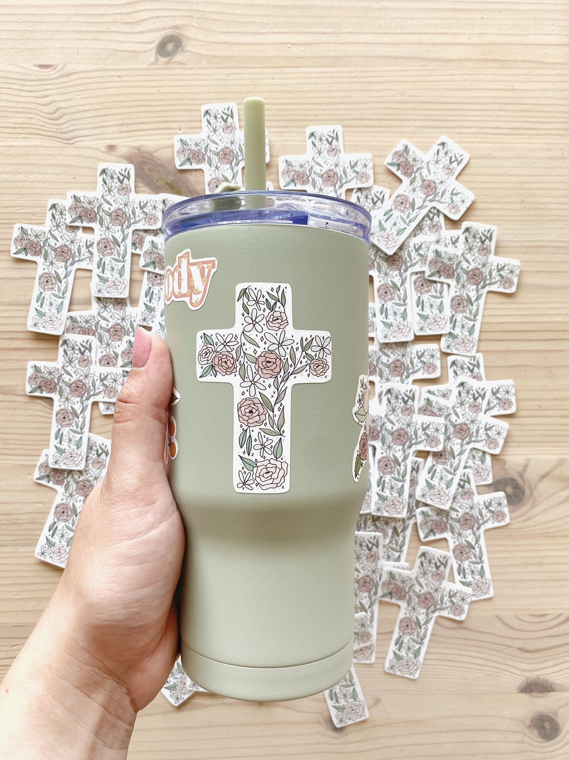 Hand holding a sage green tumbler with a floral cross sticker, with matching stickers spread out in the background.