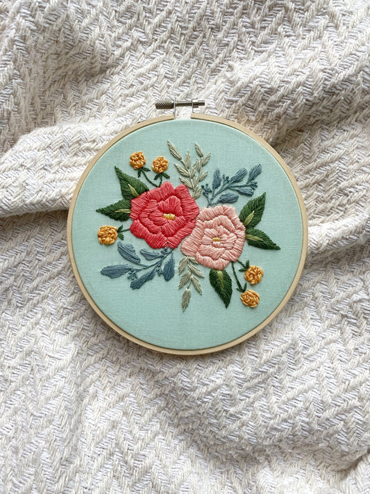 A 6-inch embroidery hoop artwork featuring a floral design with two large peonies, greenery, and sweet buds or billy ball flowers in a green background, framed in a light-colored embroidery hoop.