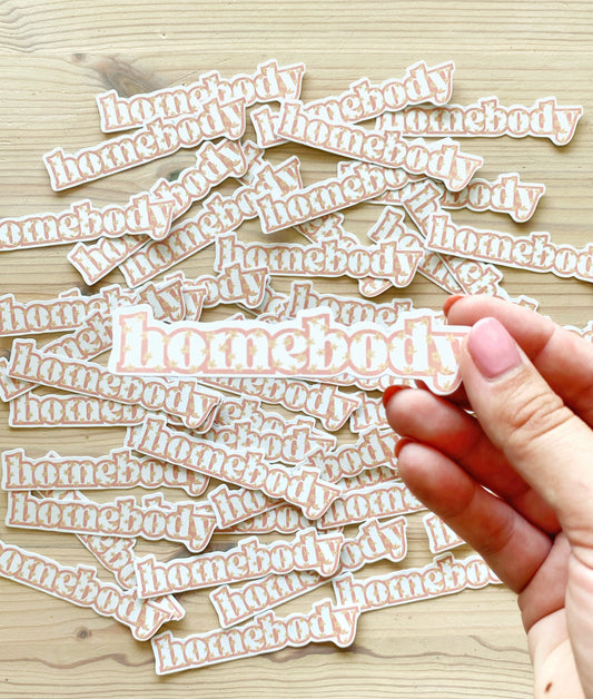 Hand holding a vinyl homebody sticker with little flowers on it.