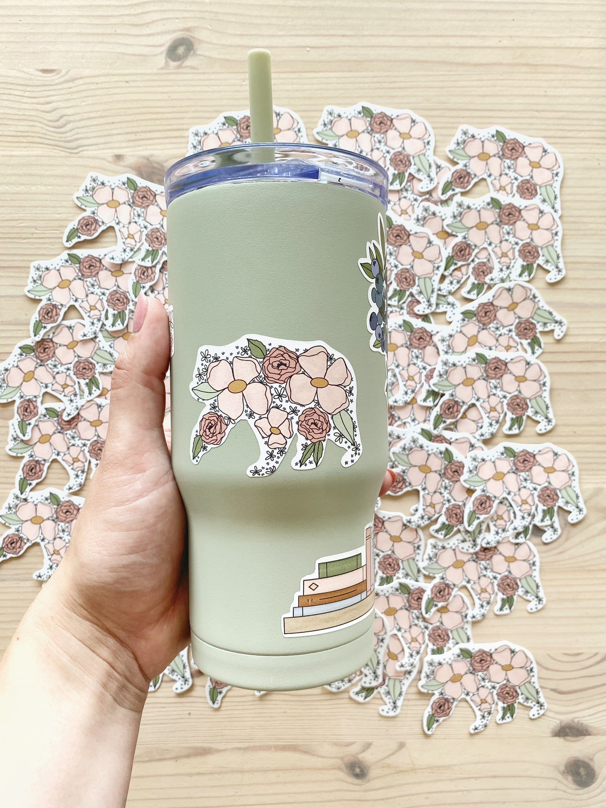 A hand holds a sage green drink tumbler decorated with a floral mama bear silhouette sticker, displayed on a background of matching mama bear stickers.