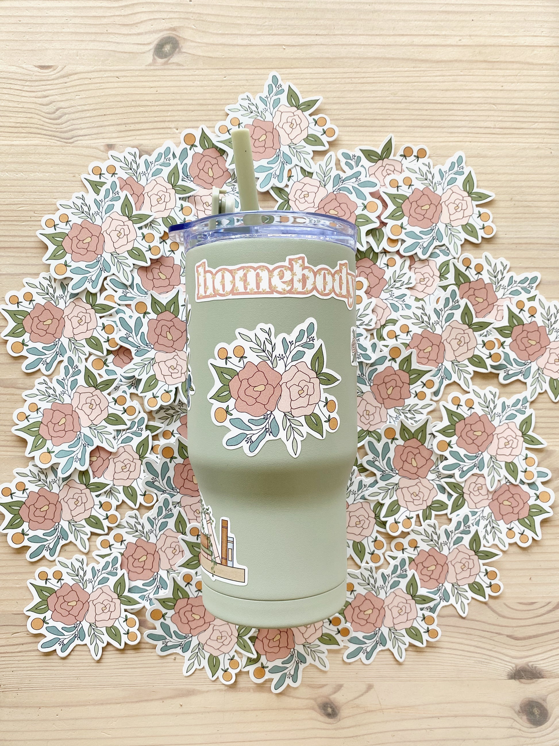 A sage green tumbler featuring a floral arrangement design sits on atop a layer of matching pink peony stickers.