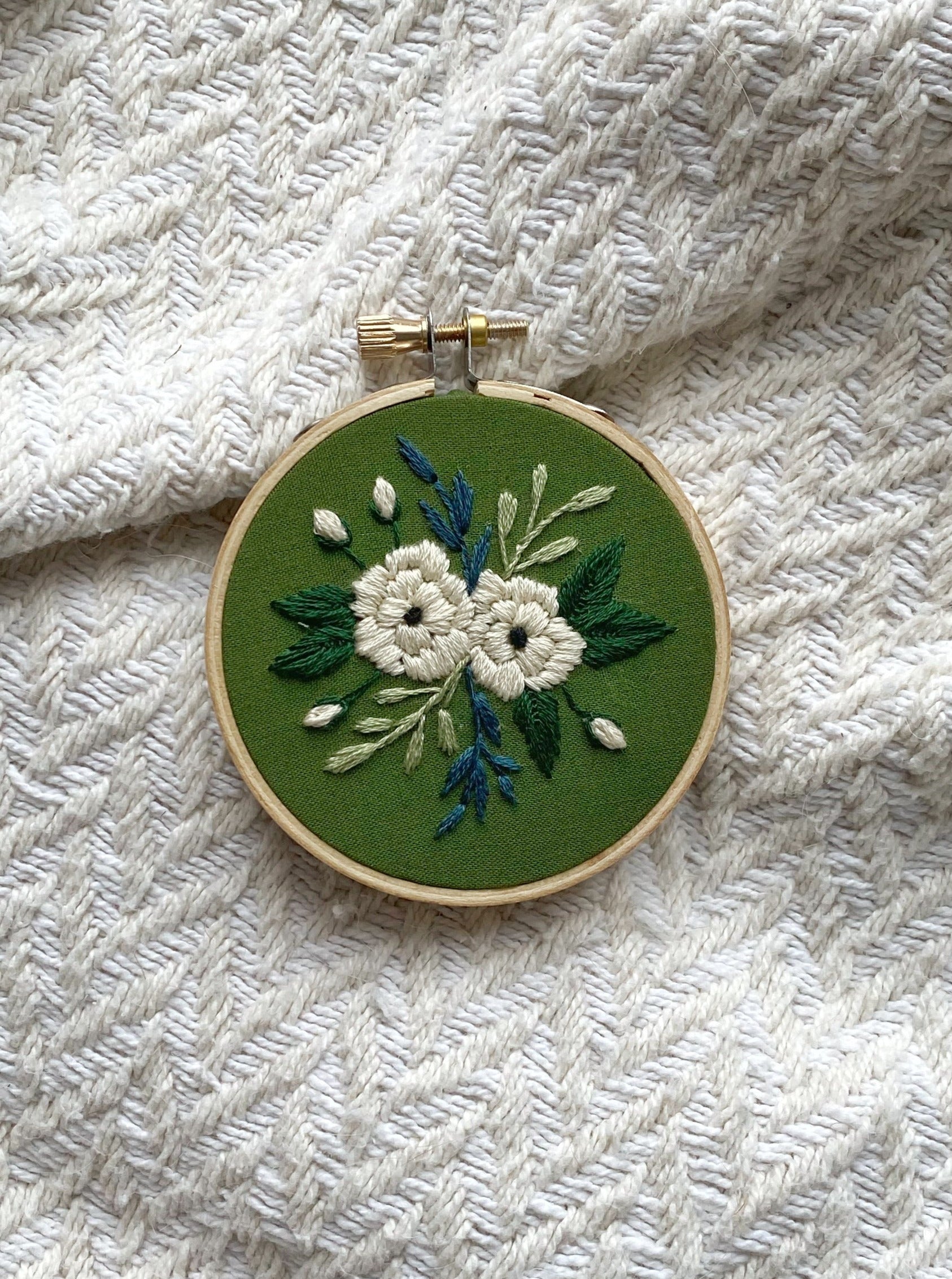 An embroidery hoop art featuring a variety of greenery and small blooms surrounding two large white peonies stitched on deep green fabric and framed in a 3 inch wooden hoop on a textured display background.