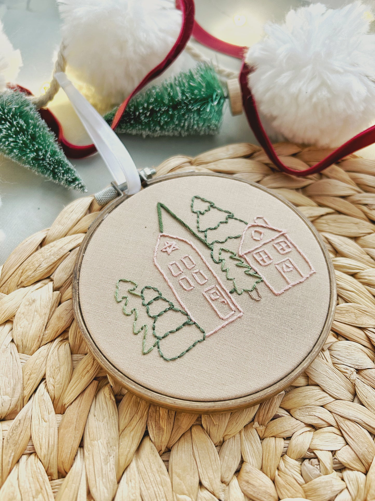 A hand embroidered holiday ornament featuring a Christmas village made by a stick and stitch kit is artfully displayed on a textured surface surrounded by seasonal decorations.