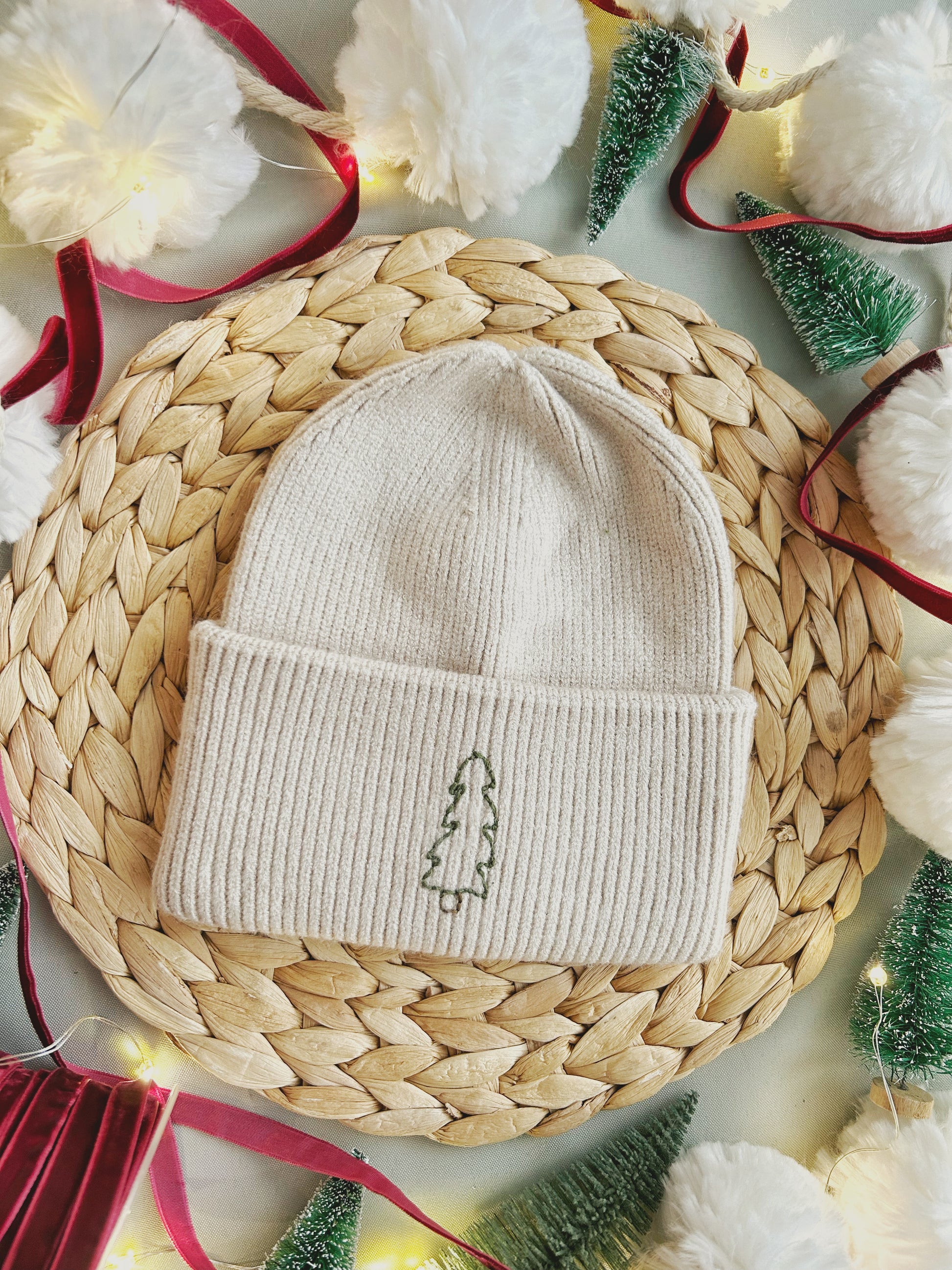 A hand embroidered toque done with a stick and stitch patch sits atop a textured surface and surrounded by small Christmas trees, garland, ribbon and twinkle lights.