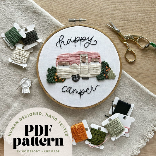 Embroidery hoop with 'Happy Camper' design, surrounded by embroidery threads and scissors on a textured fabric background. Text overlay on image denotes "hand tested, human designed PDF pattern by Homebody Handmade"