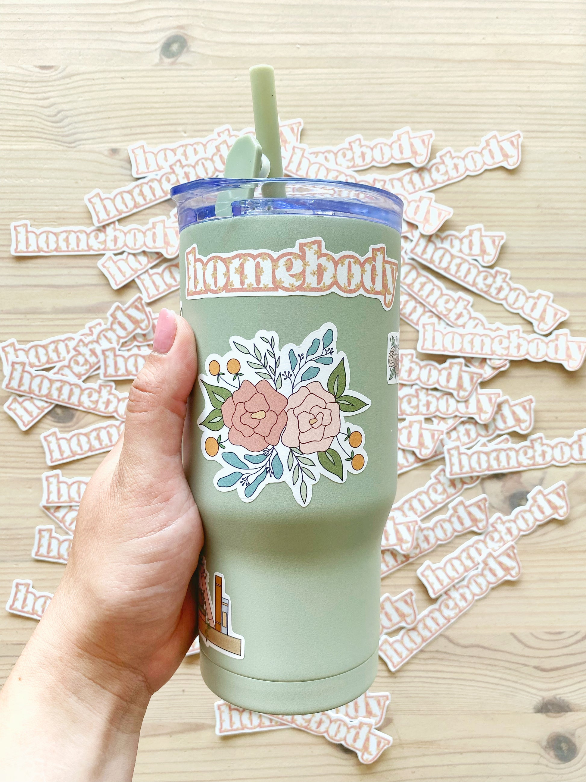 Hand holding a light green tumbler with floral and ‘homebody’ stickers, matching homebody stickers in the background.