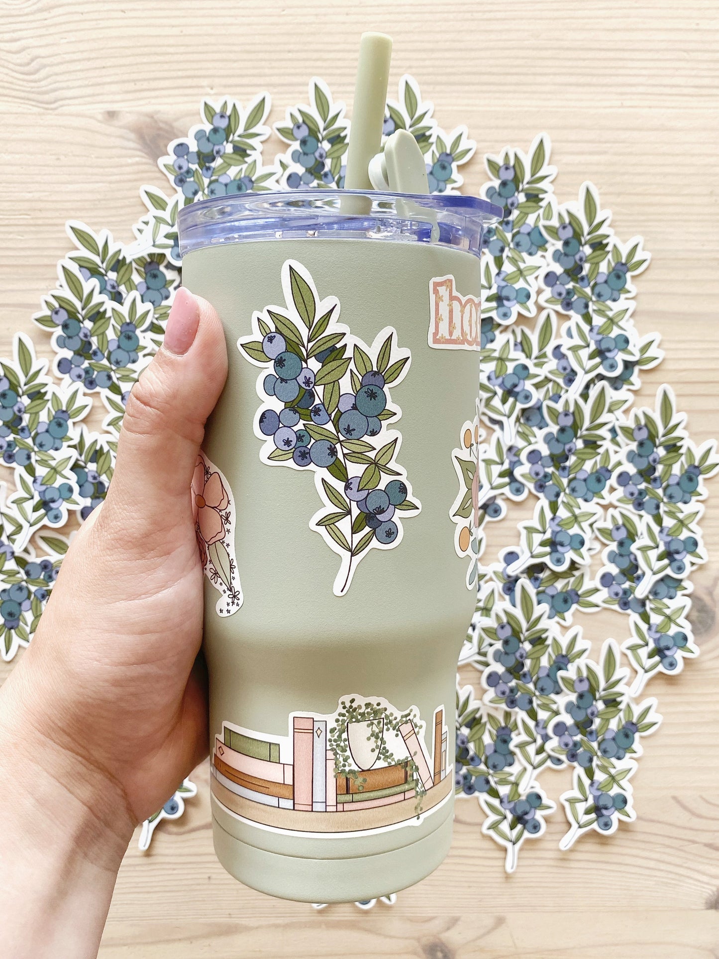 A hand holds a sage green tumbler water bottle featuring illustrated stickers - one depicting blueberries and another of a bookshelf.