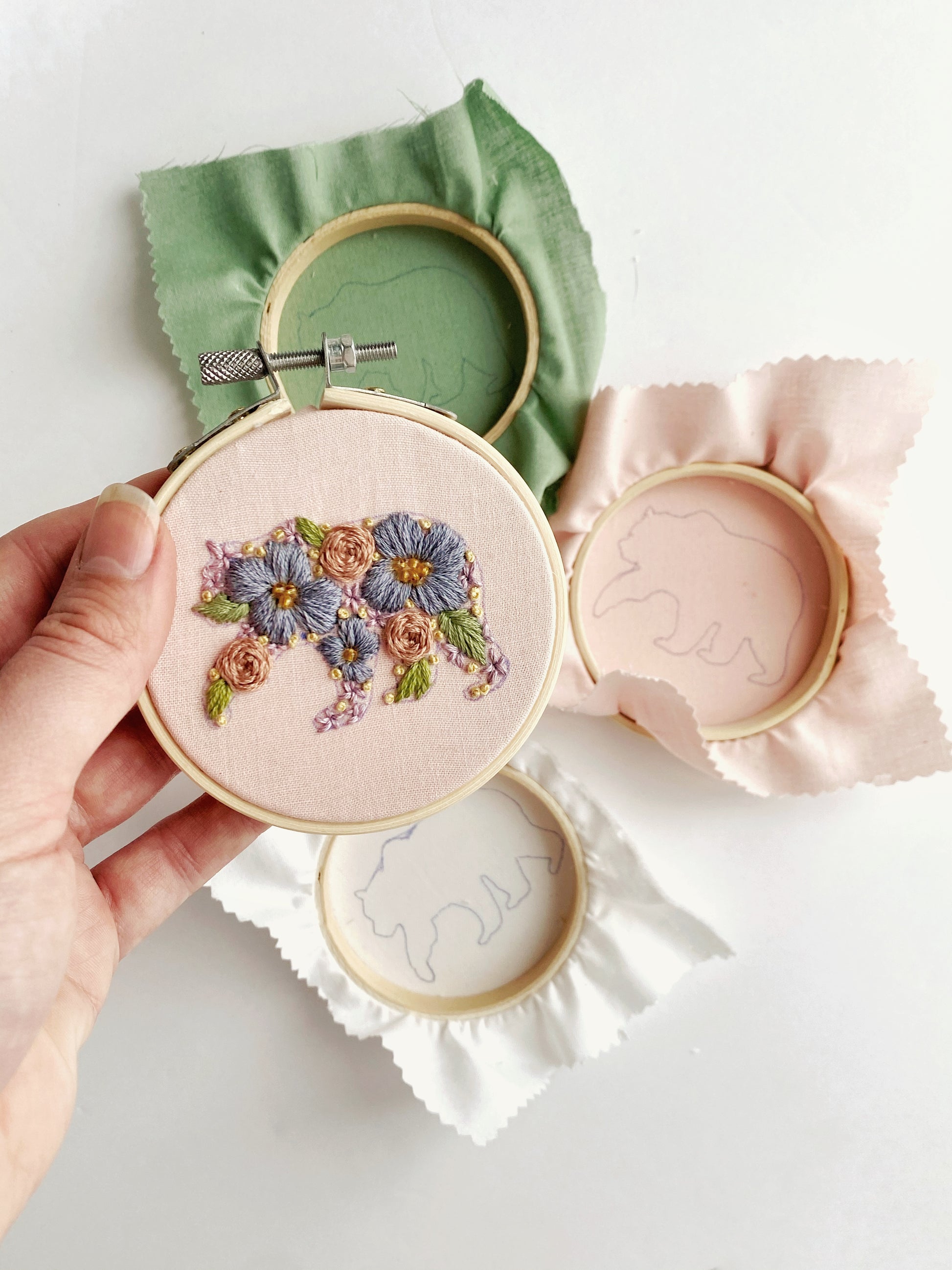 A hand holds a small embroider hoop featuring a bear silhouette filled in entirely by flowers with multiple hoops showing the pattern are in the background.