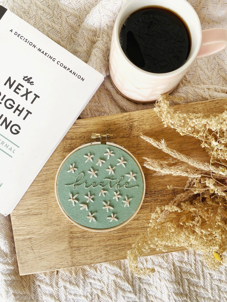 A finished embroidery hoop featuring the word breathe stitched in cursive in green thread on a matching fabric background. Small daisies fill in the blank space around the rest of the fabric. The hoop sits on a wood plank with a textured background with a coffee and a book. 