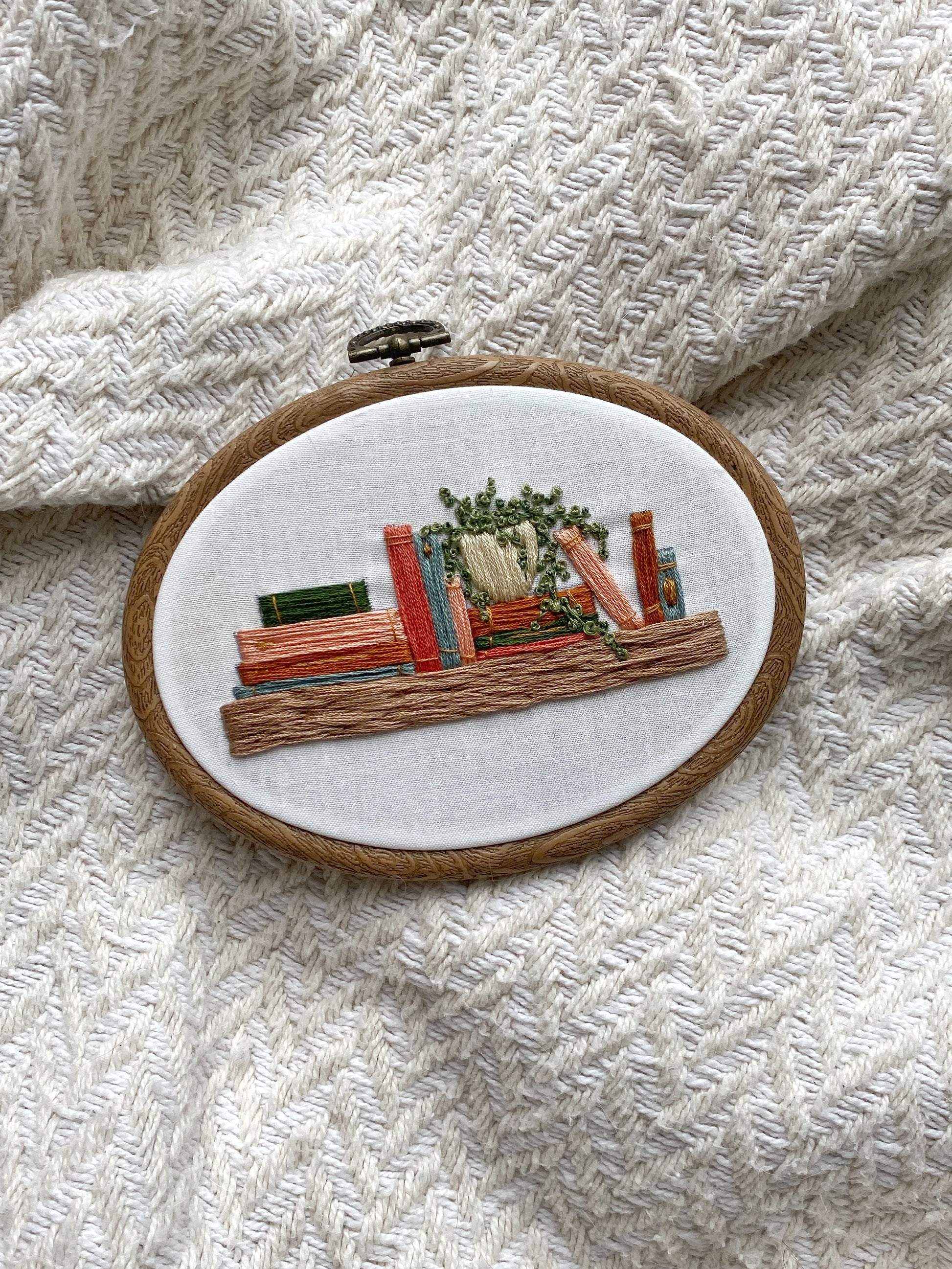 An embroidery hoop featuring a bookshelf with a potted plant for embroidery design inspiration.
