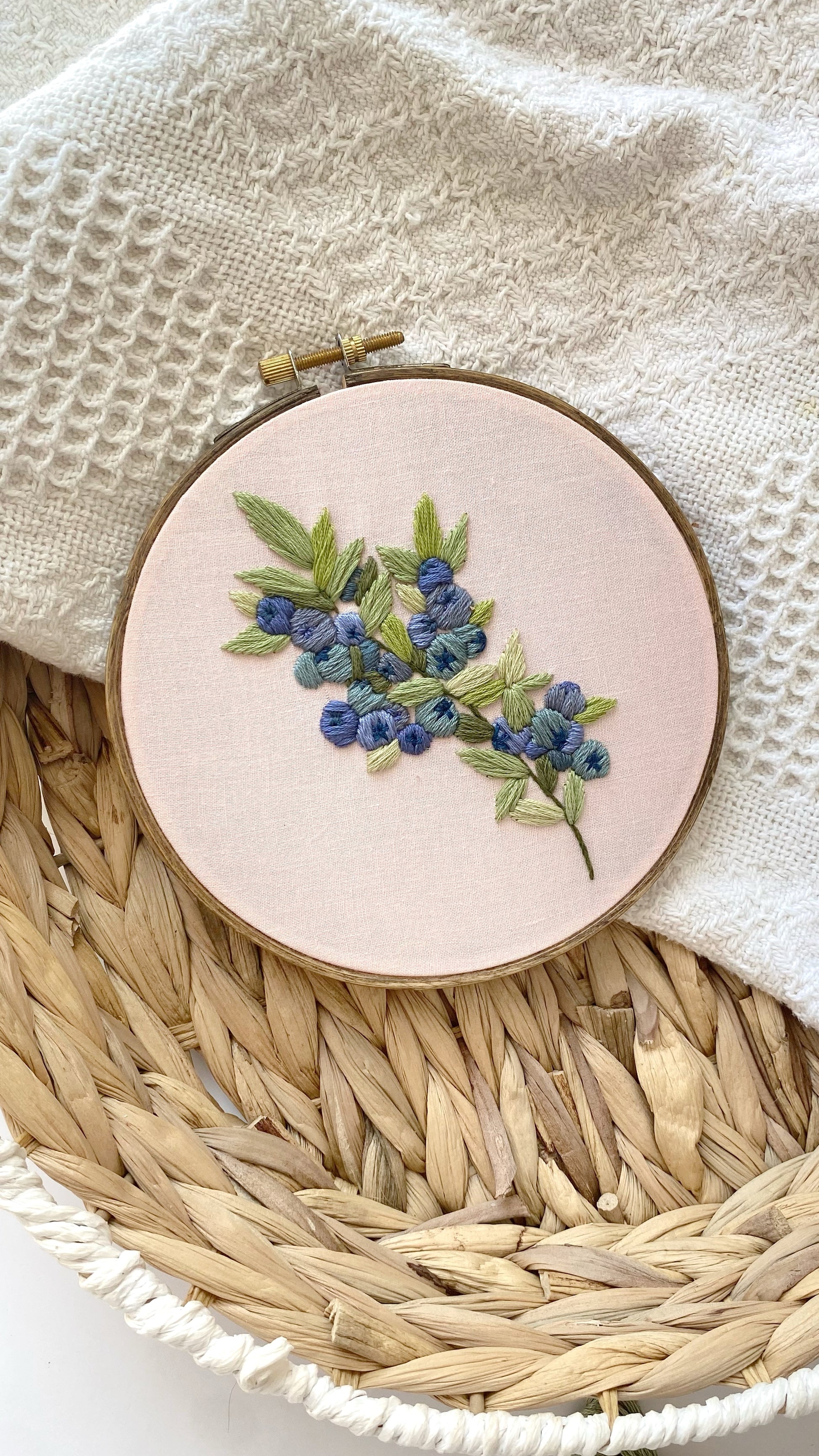 Embroidery hoop with blueberry design on a light pink fabric, placed on a woven surface.