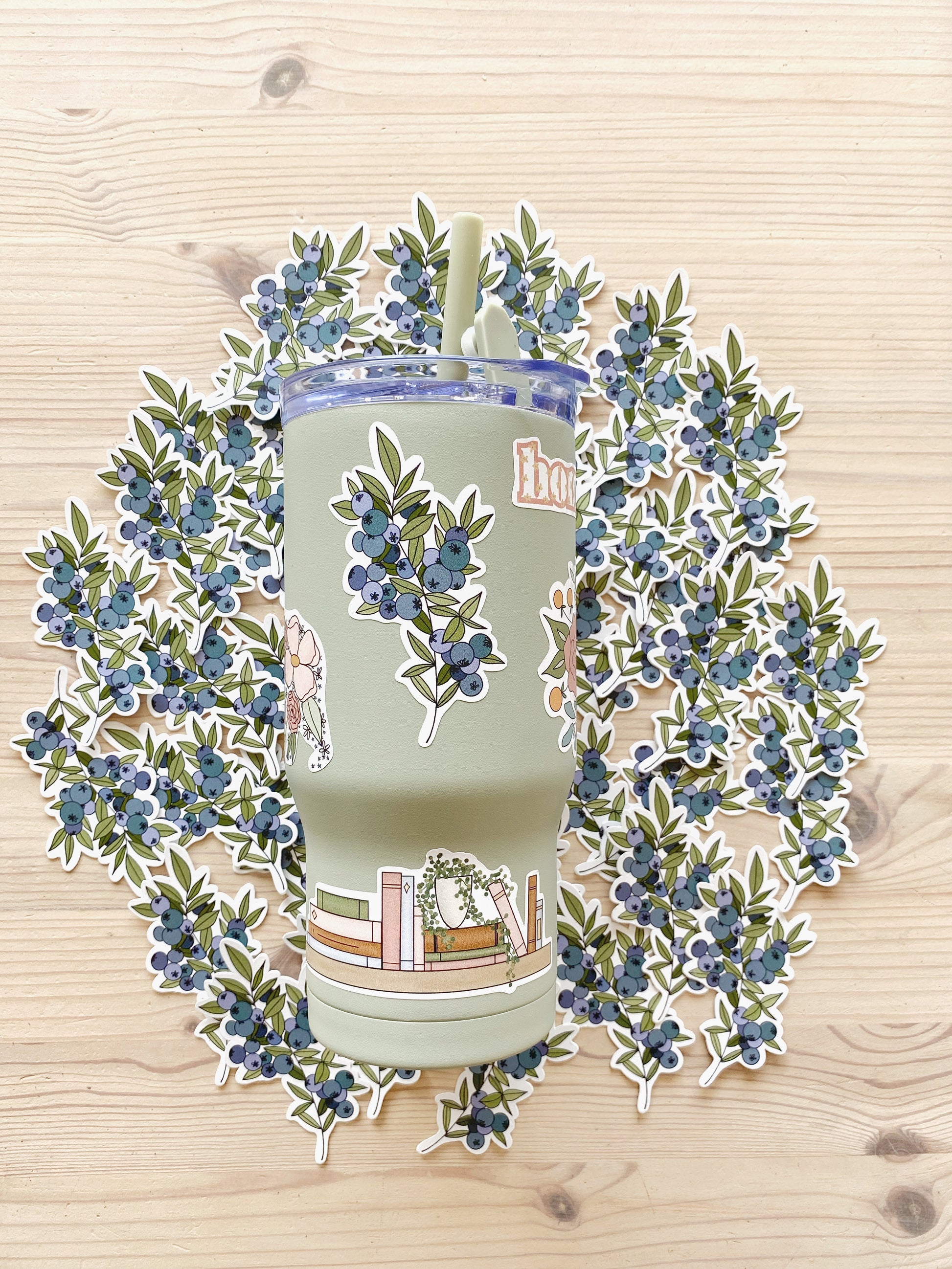 Sage green cup adorned with multiple illustrated stickers including a blueberry sprig and a bookshelf with a potted plant sits atop more blueberry stickers in the background. 