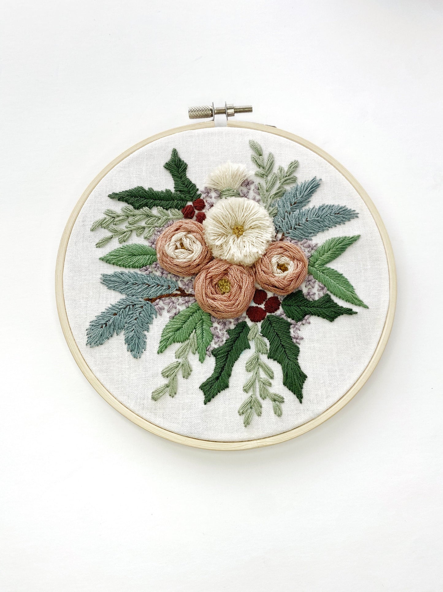 A large arrangement of embroidered flowers featuring pink boughs, holly, and other greenery with textured roses and fluffy flowers framed in a 6 inch embroidery hoop.