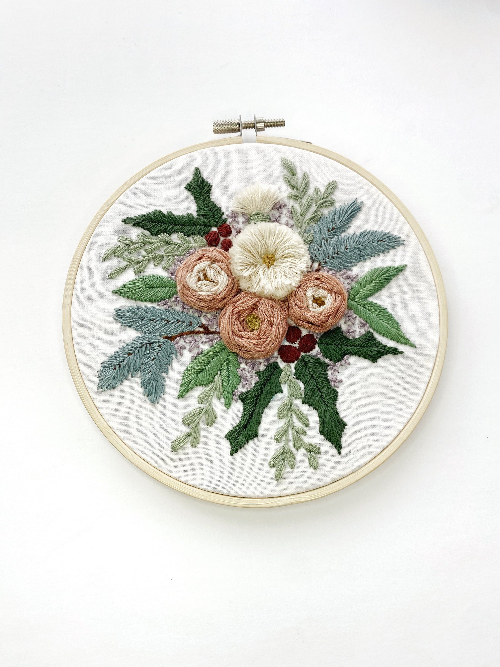 A large arrangement of embroidered flowers featuring pink boughs, holly, and other greenery with textured roses and fluffy flowers framed in a 6 inch embroidery hoop.