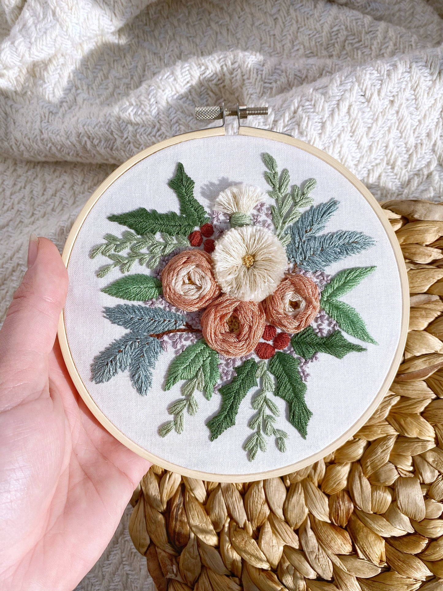 An arrangement of winter foliage including pine and holly with textured roses and other flowers is embroidered on white fabric and framed in a 6 inch hoop.